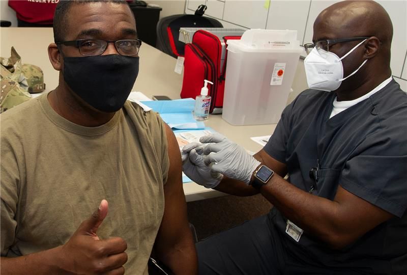 A man wearing a black face mask and glasses gives a thumbs-up while receiving a vaccination shot in his upper arm from a healthcare worker wearing a white face mask, gloves, and dark scrubs. They are in a clinical setting with medical supplies on a table behind them.